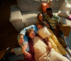 Four friends lounging together on a large corduroy sofa in a sunlit living room captured from a high-angle overhead perspective.