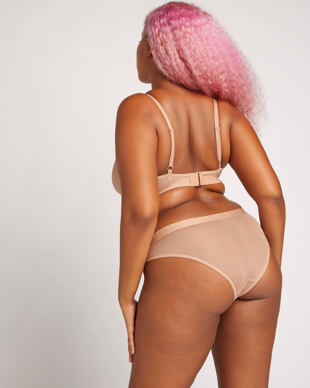 Back view of an adult woman with pink hair in a beige lingerie set standing in a white studio with soft lighting. Fashion or beauty shot.