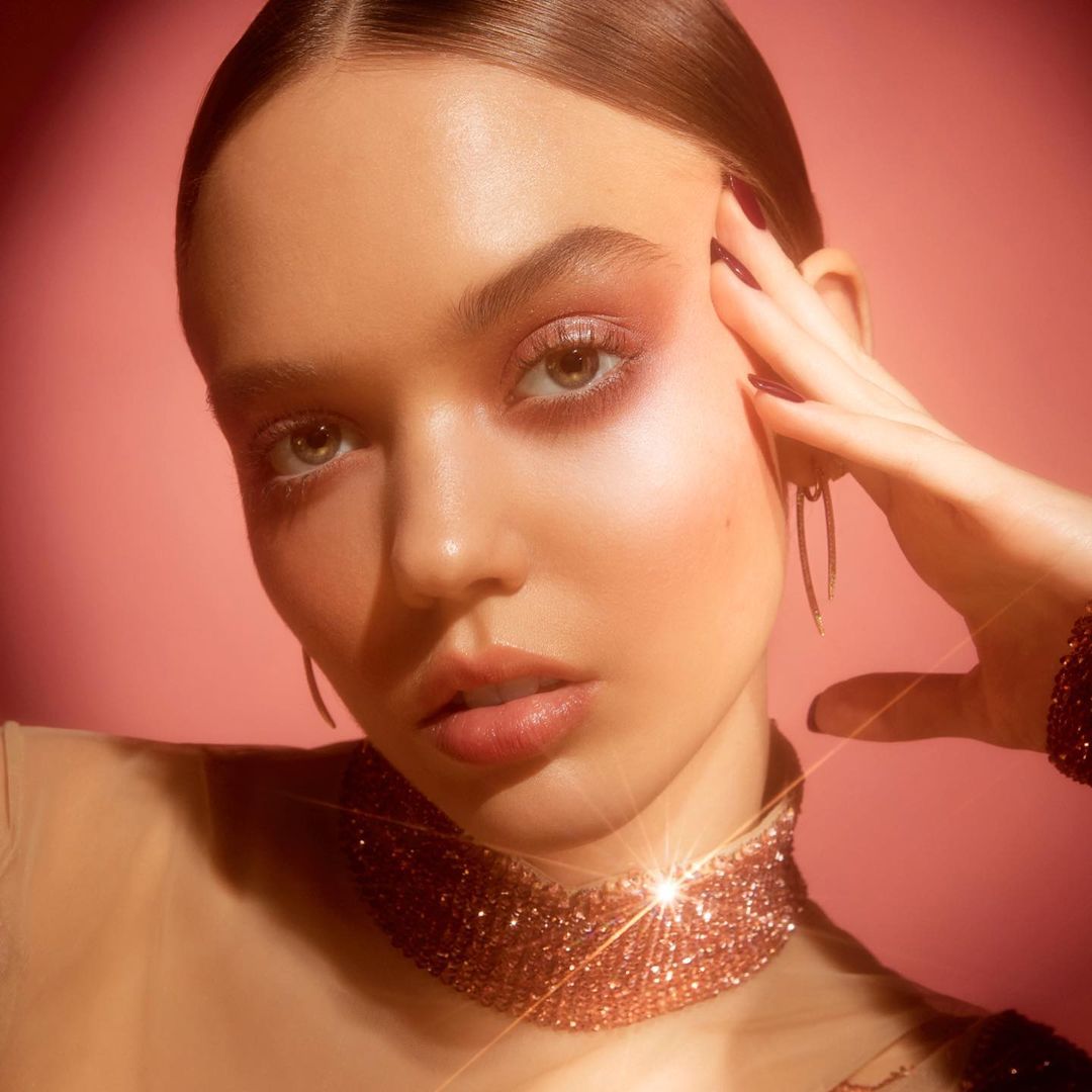Close-up portrait of a woman with pink eyeshadow, glossy lips, a shimmering choker, and styled hair against a pink background.