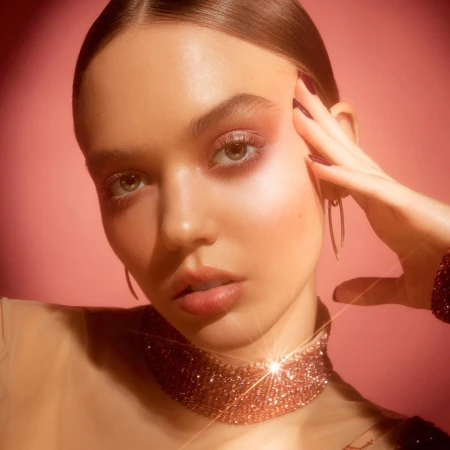 Close-up portrait of a woman with pink eyeshadow, glossy lips, a shimmering choker, and styled hair against a pink background.