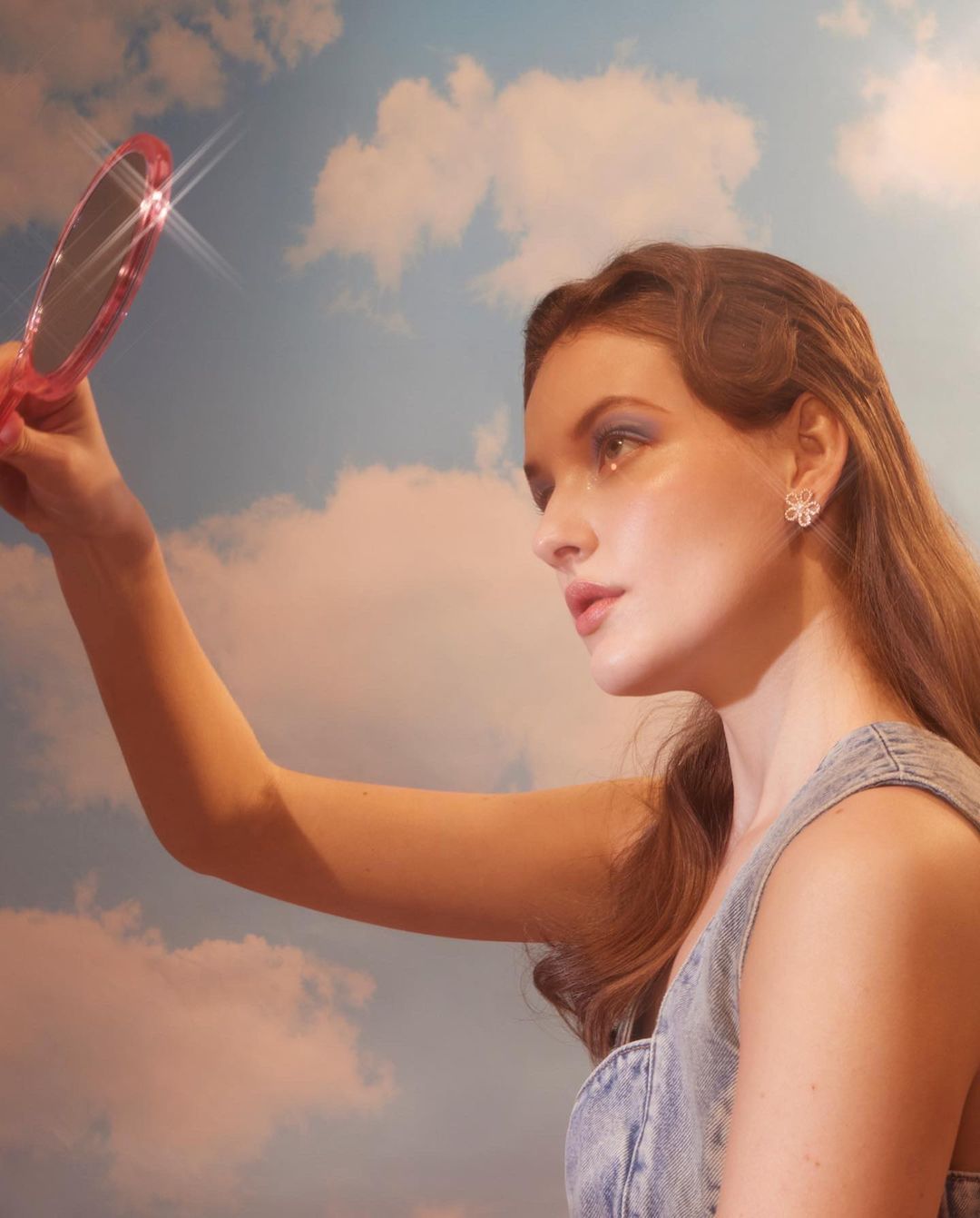 Young woman wearing a denim dress and flower earrings holds a hand mirror, posing against a backdrop of blue sky and white clouds.