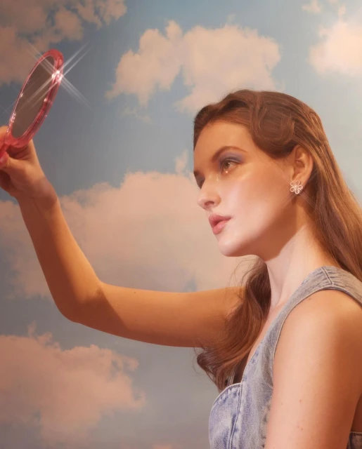 Young woman wearing a denim dress and flower earrings holds a hand mirror, posing against a backdrop of blue sky and white clouds.