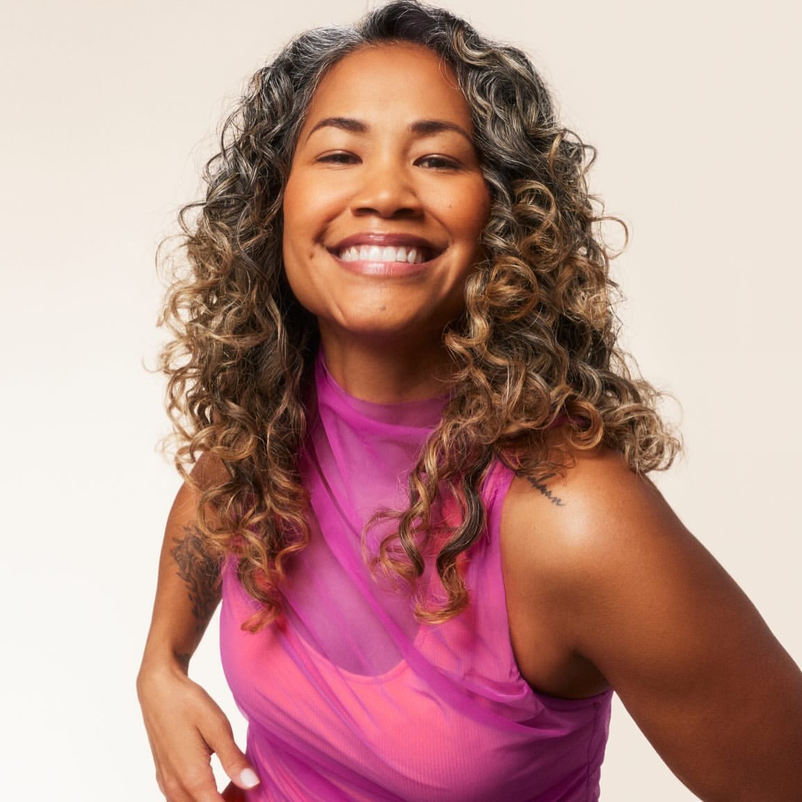 Close-up of a smiling mixed-race woman with curly gray-streaked hair, wearing a bright pink top against a neutral background.