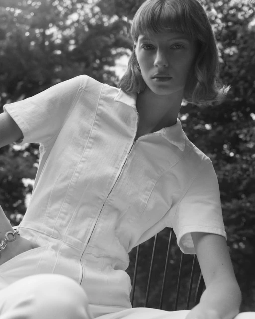 Portrait of a young woman with bangs in a white jumpsuit sitting in a chair outdoors with trees visible in the background in black and white.