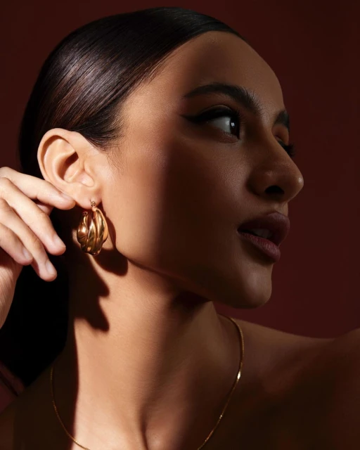 Close-up of a woman with dark hair and winged eyeliner wearing gold hoop earrings and a gold chain against a dark red background.