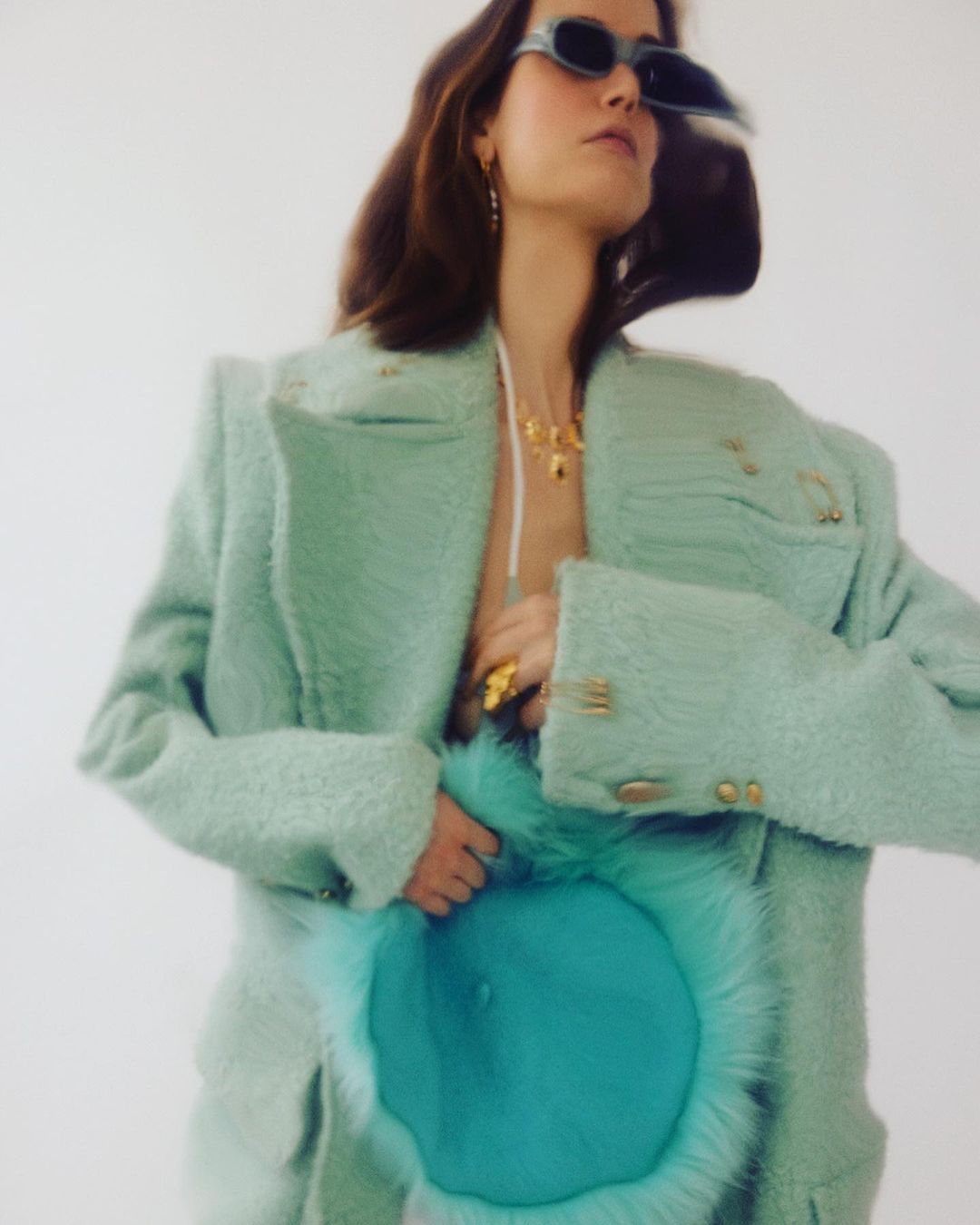Adult woman wearing a green tweed coat and sunglasses holding a furry blue handbag, posing in front of a white backdrop.
