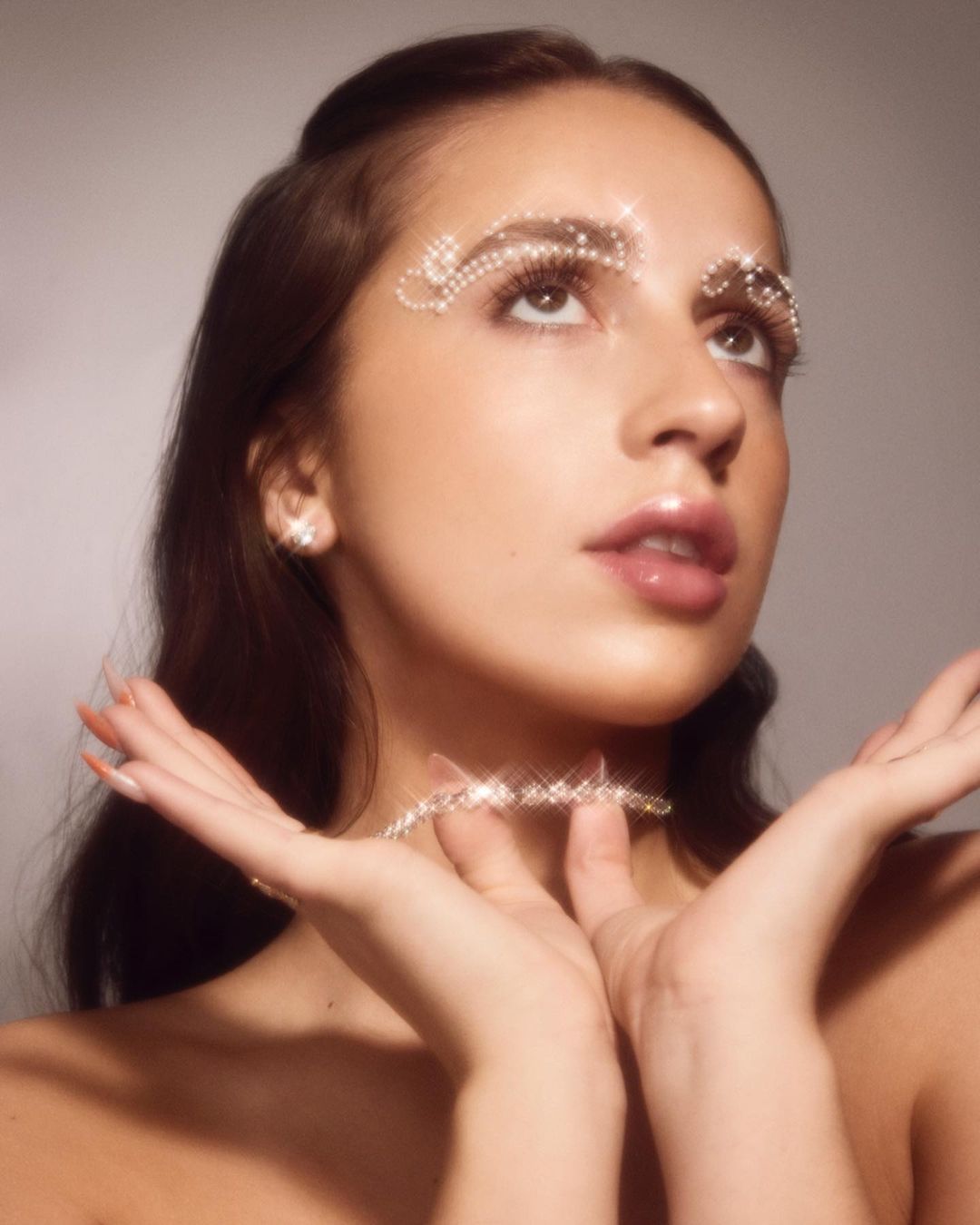 Close-up portrait of a young woman wearing decorative jewelry around her eyes and neck. She is looking up with her hands raised.