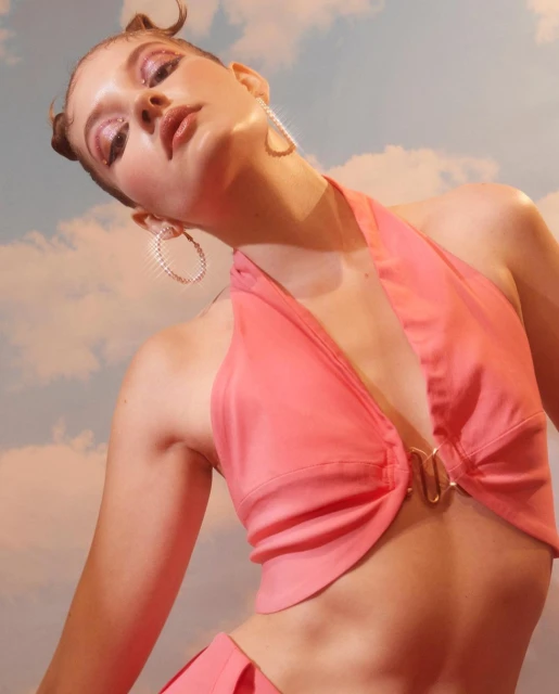 Young adult woman poses in a pink halter top with gold hardware, hoop earrings, and space buns, against a blue and white cloudy sky background.