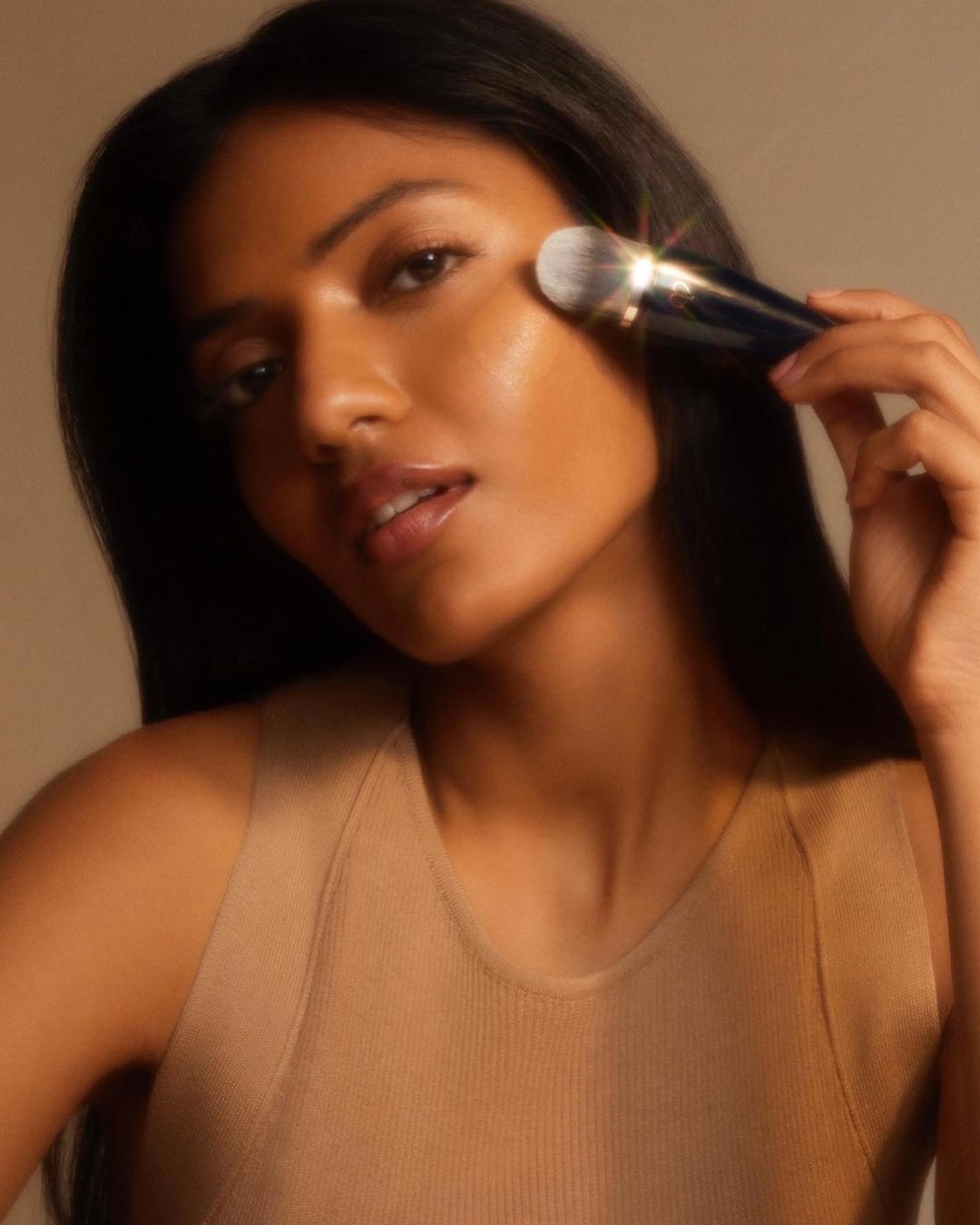 Elegant portrait of a dark-haired woman applying makeup with a brush to her face in a softly lit beauty or fashion context.