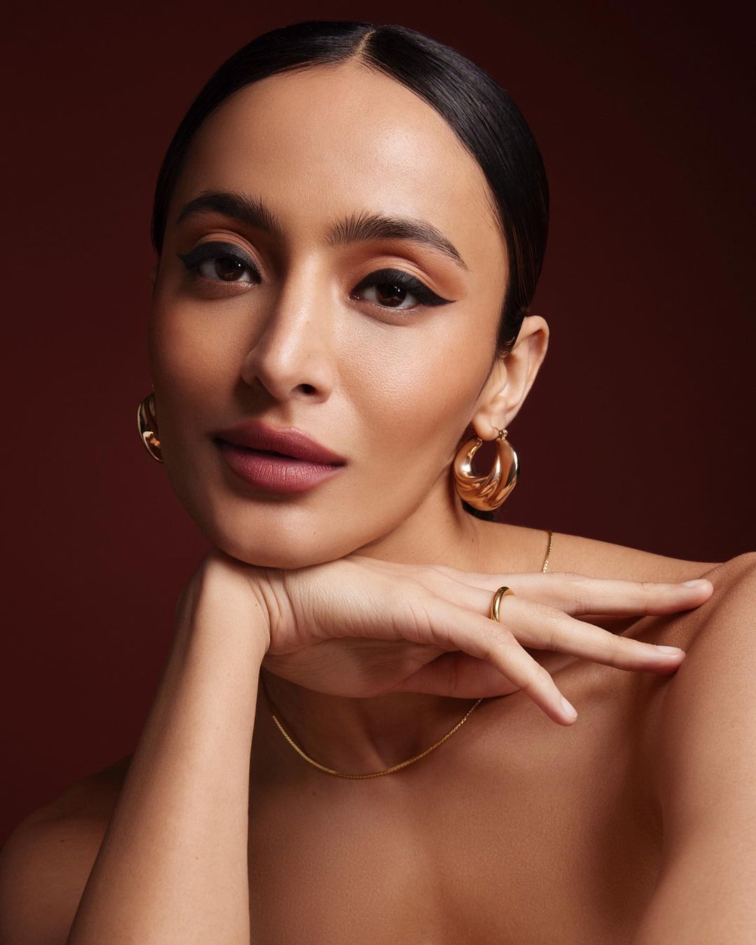 Portrait of a woman with her face resting on her hand, wearing gold hoop earrings and a gold necklace, set against a brown backdrop.