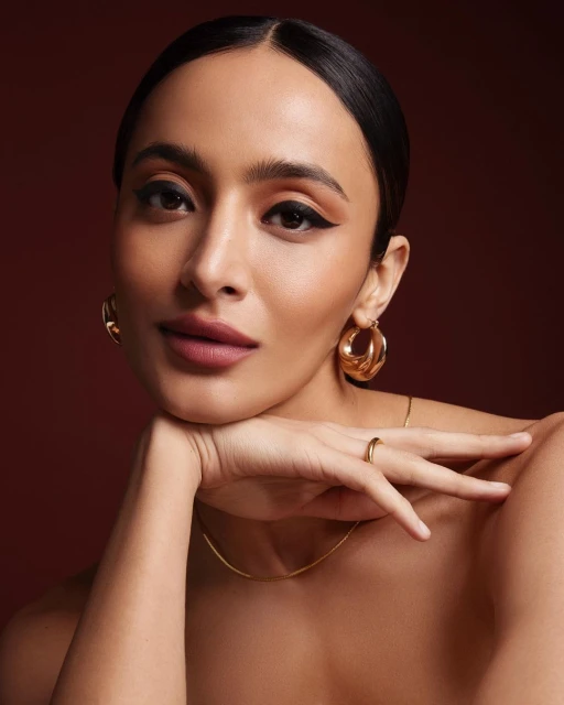 Portrait of a woman with her face resting on her hand, wearing gold hoop earrings and a gold necklace, set against a brown backdrop.