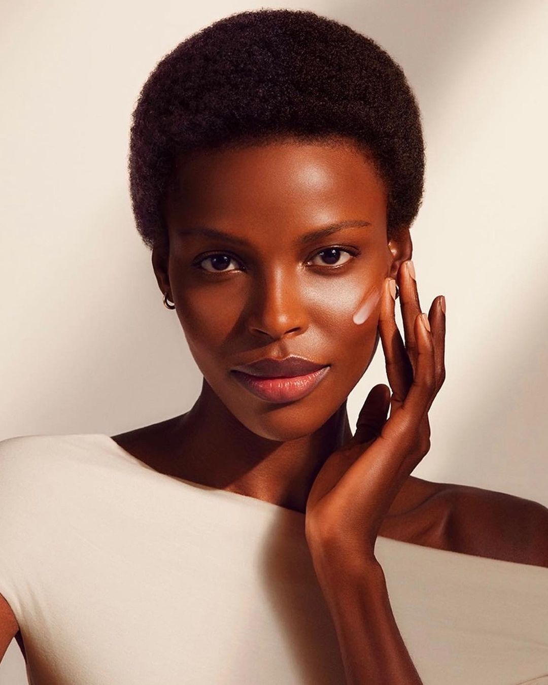Close-up of a Black woman applying skincare product to her face, wearing an off-white top, in a beauty studio with soft lighting.