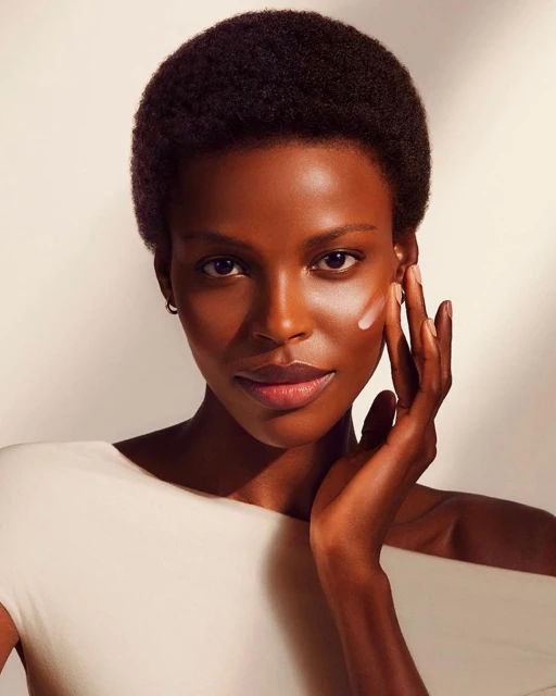 Close-up of a Black woman applying skincare product to her face, wearing an off-white top, in a beauty studio with soft lighting.
