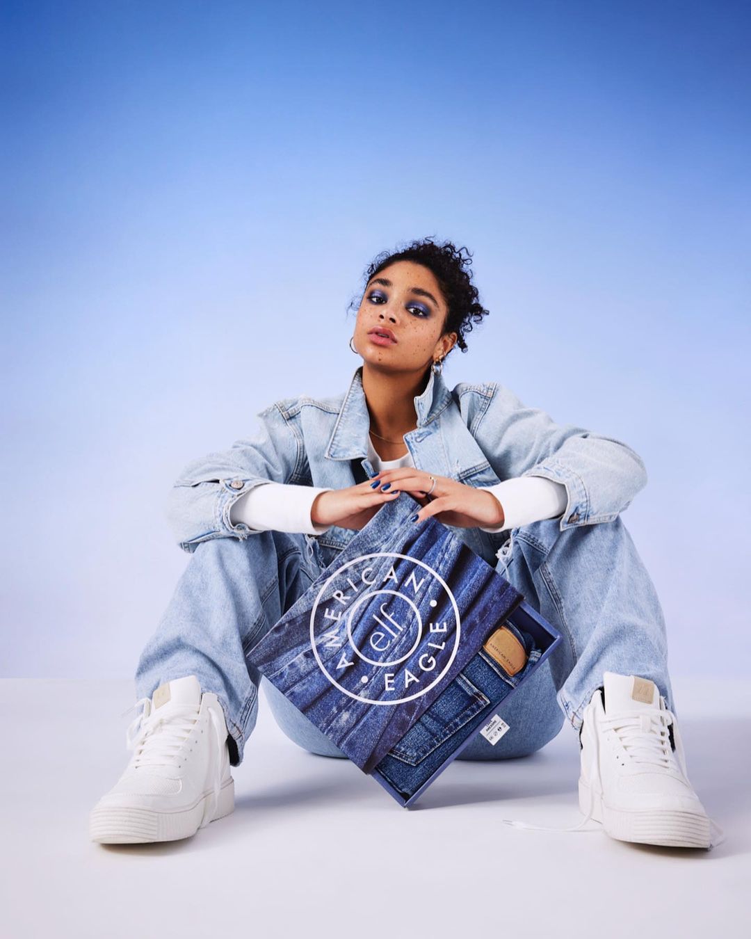Young woman in casual denim outfit poses with an American Eagle denim gift box in a studio with a light blue gradient background.
