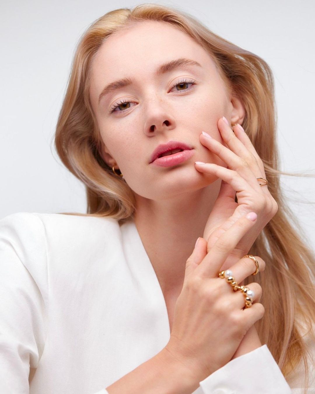 Close-up of a fair-skinned woman with blond hair in a white jacket, showcasing gold rings on her hand against a white backdrop.