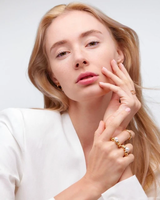 Close-up of a fair-skinned woman with blond hair in a white jacket, showcasing gold rings on her hand against a white backdrop.