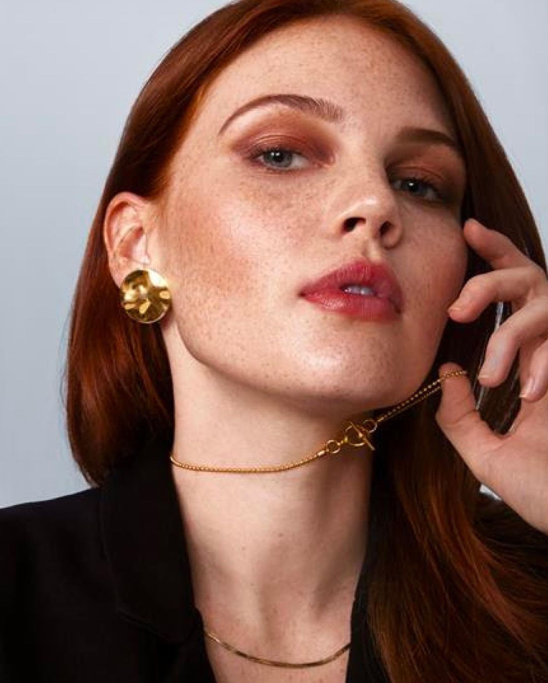 Close-up of a red-haired woman indoors wearing a black blazer and gold jewelry: earrings and a toggle clasp necklace.