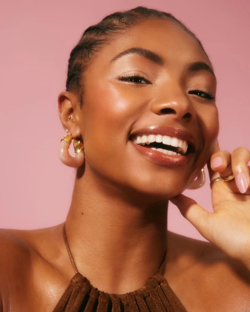 Close-up of a smiling Black woman with hoop earrings and brown halter top against a pink background in a beauty advertising shot.