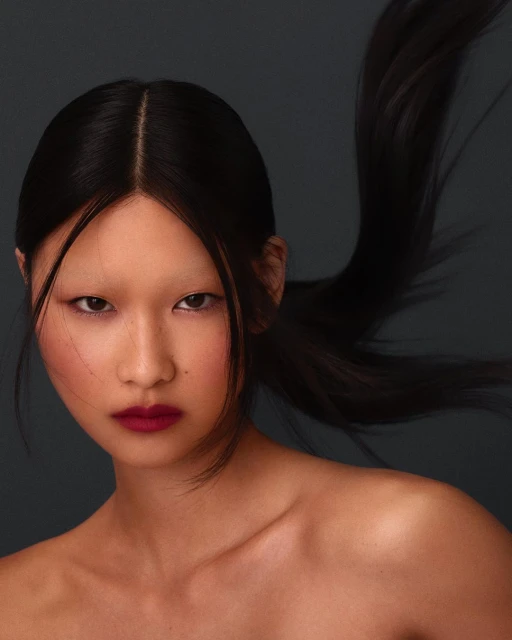 Portrait of a young Asian woman with dark hair slicked back, dramatic red lipstick, and pale eyebrows against a dark gray backdrop.