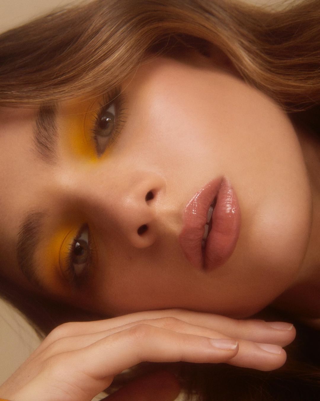 Close-up portrait of a young adult female model with yellow eyeshadow and glossy lips looking directly at the camera.