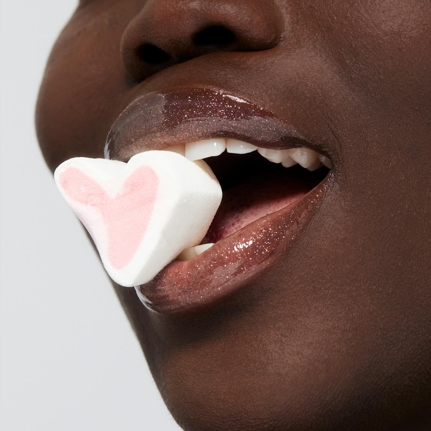 Close-up of a black woman's face holding a heart-shaped marshmallow between her teeth. The focus highlights glossy lips and the marshmallow.