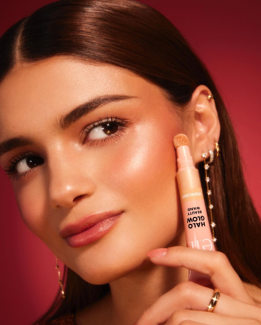 Woman applying e.l.f. Halo Glow Beauty Wand highlighter with a sponge applicator to her cheek, showcasing radiant skin and beauty product.