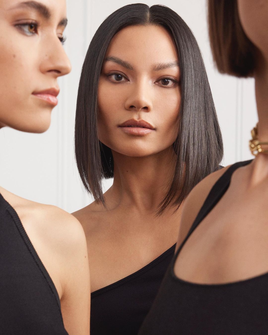 Three adult female models with dark hair pose indoors wearing black tops in a close-up studio shot, showcasing beauty and fashion.
