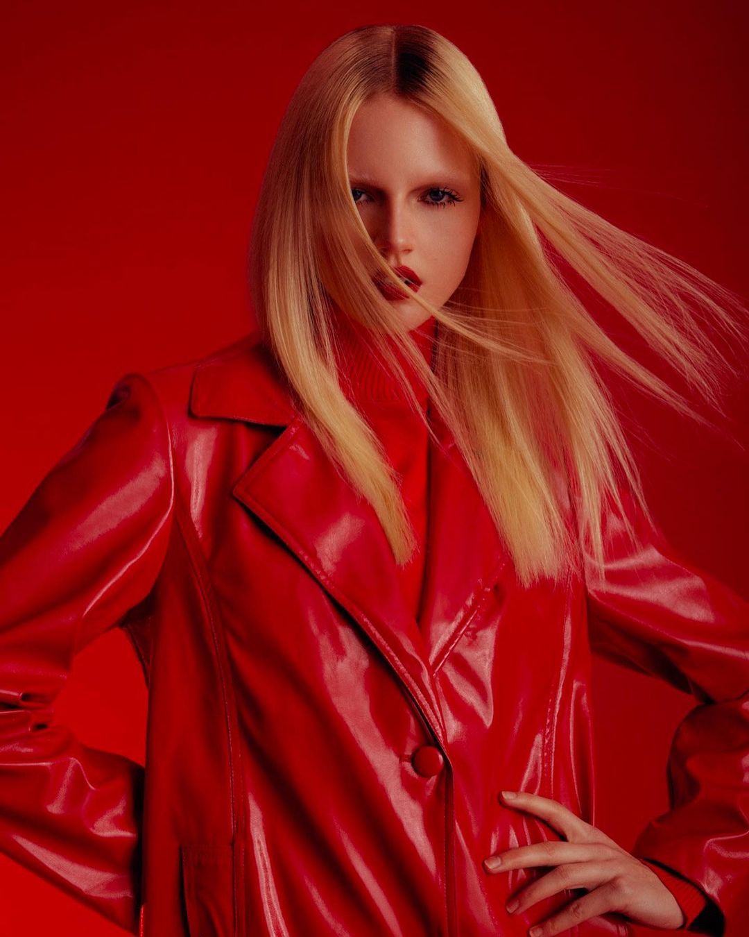 Woman with blond hair poses in a shiny red coat against a red background, fashion photography in a studio setting.