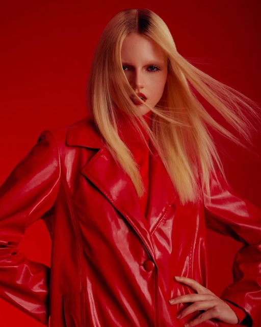 Woman with blond hair poses in a shiny red coat against a red background, fashion photography in a studio setting.