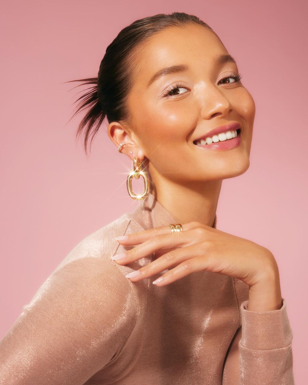 Smiling adult woman with golden jewelry poses in a studio with a pink background. She wears a sparkling beige top and ring.