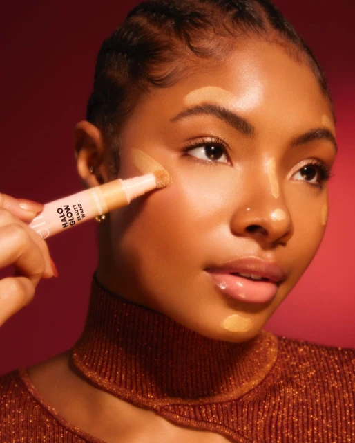 Young woman applies a light foundation to her face with a concealer stick in a beauty shot against a deep red background.