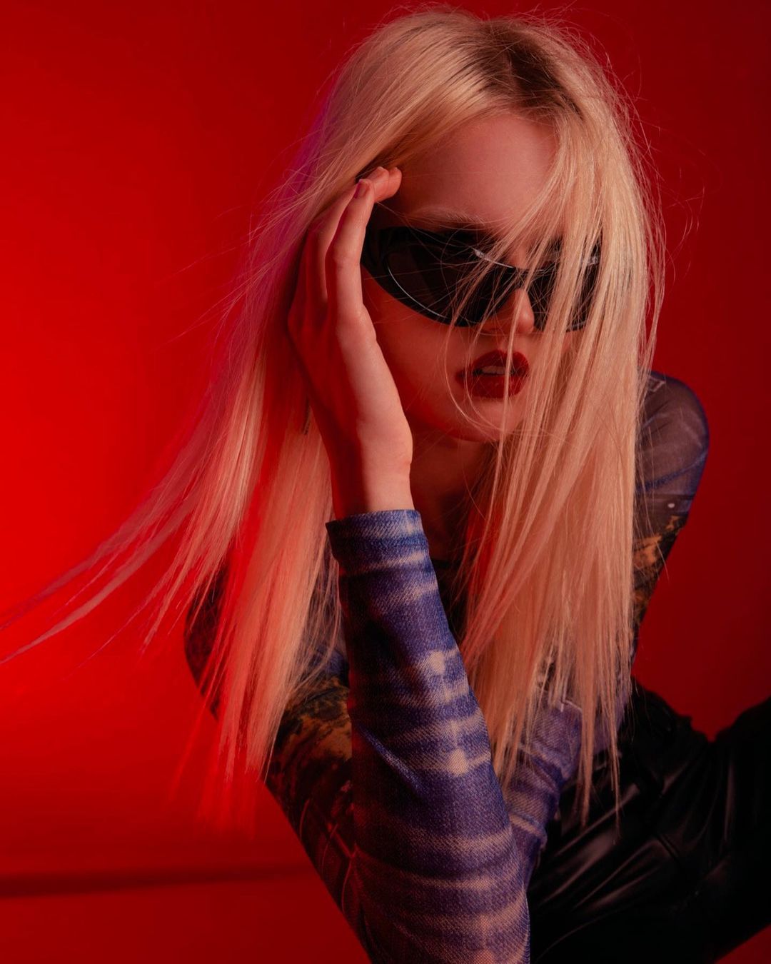 Blonde woman with sunglasses poses against a vibrant red background, showcasing a fashion-forward look and edgy style in a studio setting.