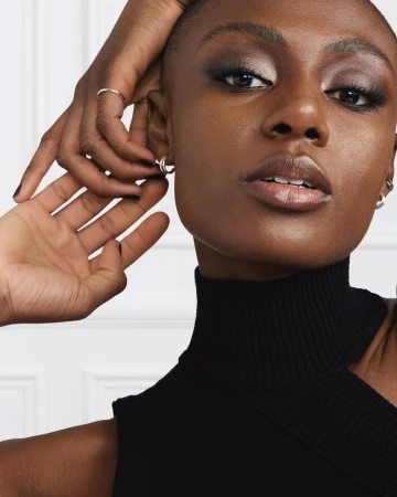Close-up of a Black woman with short hair, wearing a black turtleneck sweater. The model wears makeup and is adorned with silver jewelry.