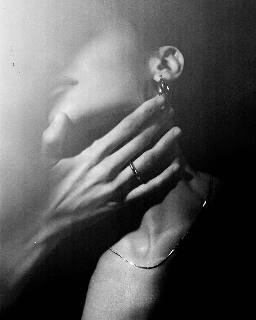 Black and white portrait featuring a woman with hands near her face, gold hoop earrings, and a delicate gold chain necklace.