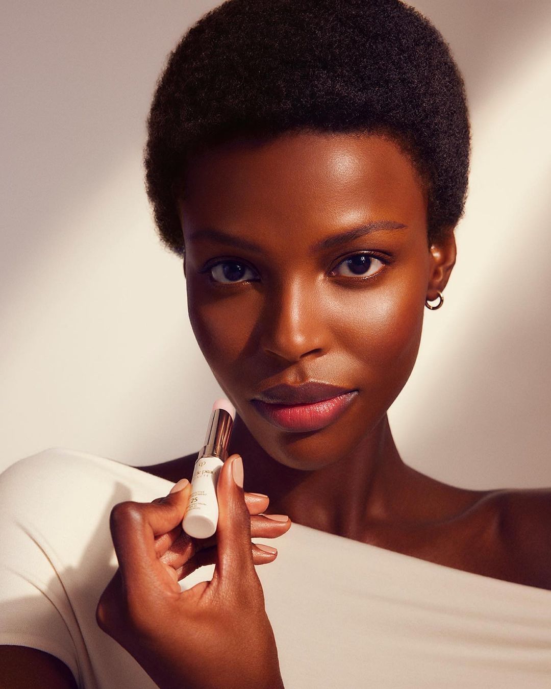 Close-up of an African American woman holding a lipstick. The model has short hair and is wearing an off-white top against a light background.