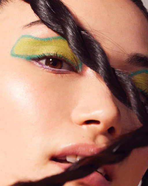 Close-up portrait of a woman with green and yellow makeup around her eyes and twisted hair framing her face.