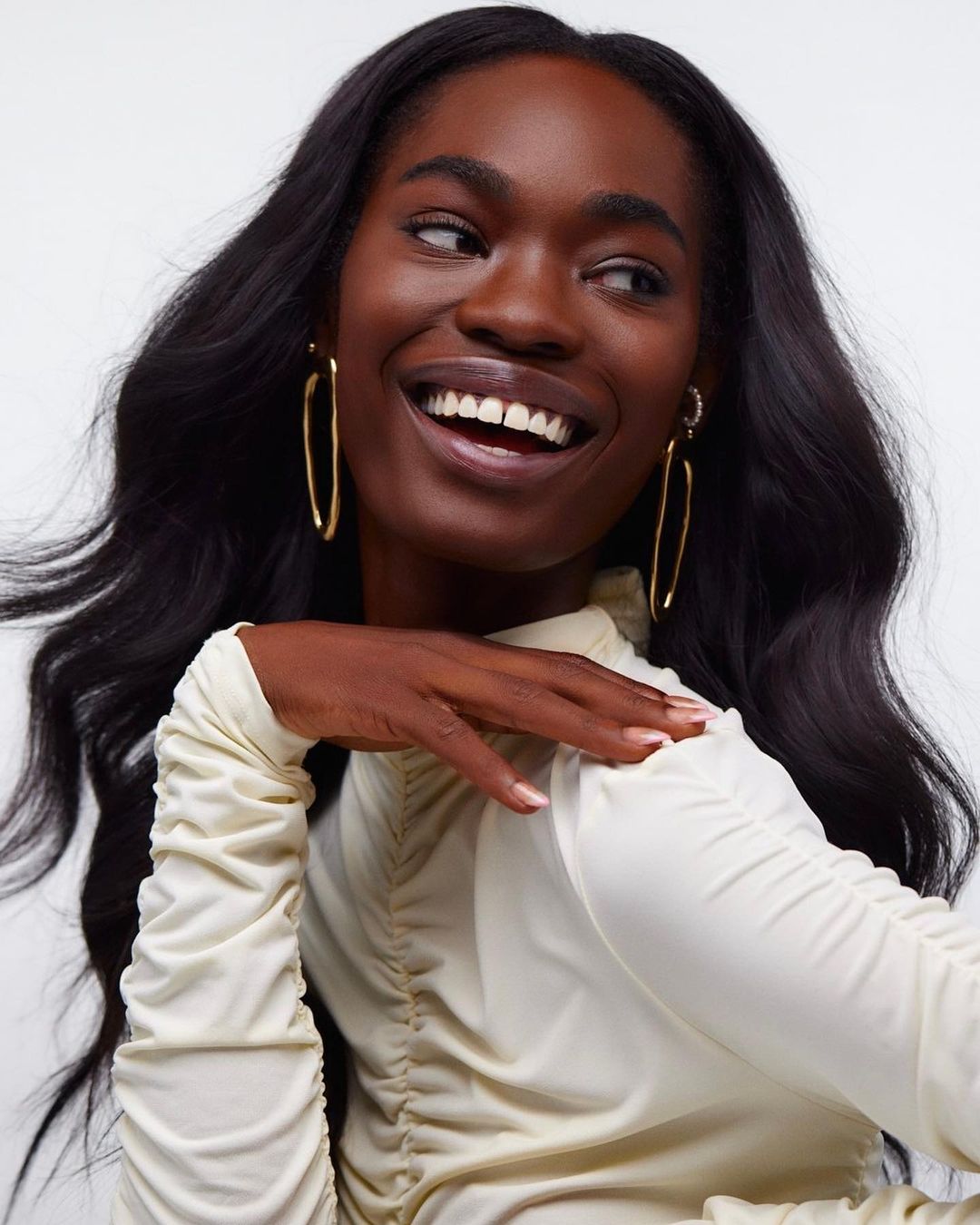 Close-up of an African American woman in a cream-colored top, smiling and wearing gold earrings, set against a plain white backdrop.
