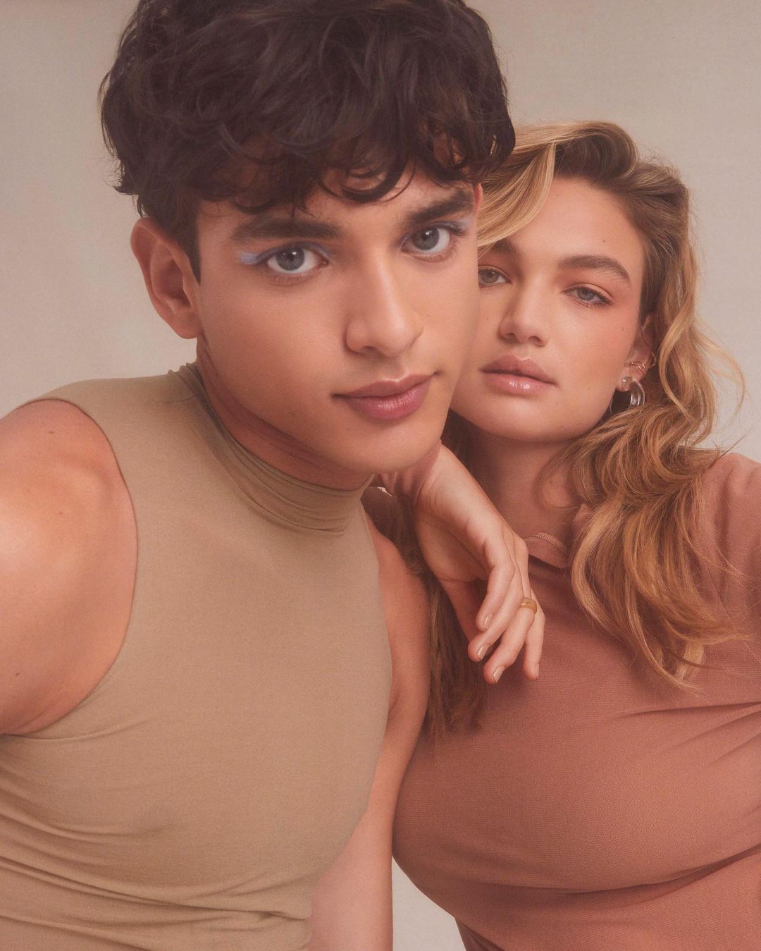Close-up of a young man and woman with makeup, standing together in a studio setting, showcasing beauty and fashion elements.
