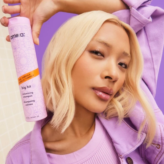 Portrait of a blonde adult woman holding a can of Amika Big Hit volumizing shampoo, wearing a lavender outfit, against a purple background.