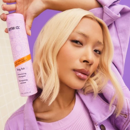 Portrait of a blonde adult woman holding a can of Amika Big Hit volumizing shampoo, wearing a lavender outfit, against a purple background.
