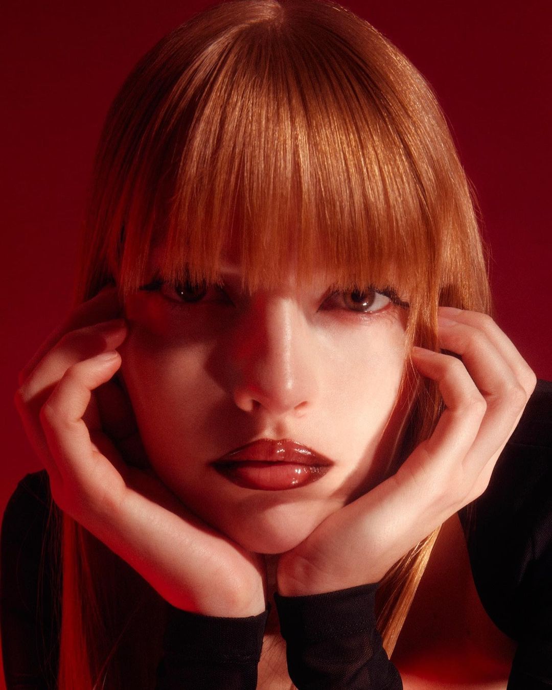 Close-up studio portrait of a young adult woman with red hair, resting her chin on her hands, set against a vibrant red background.