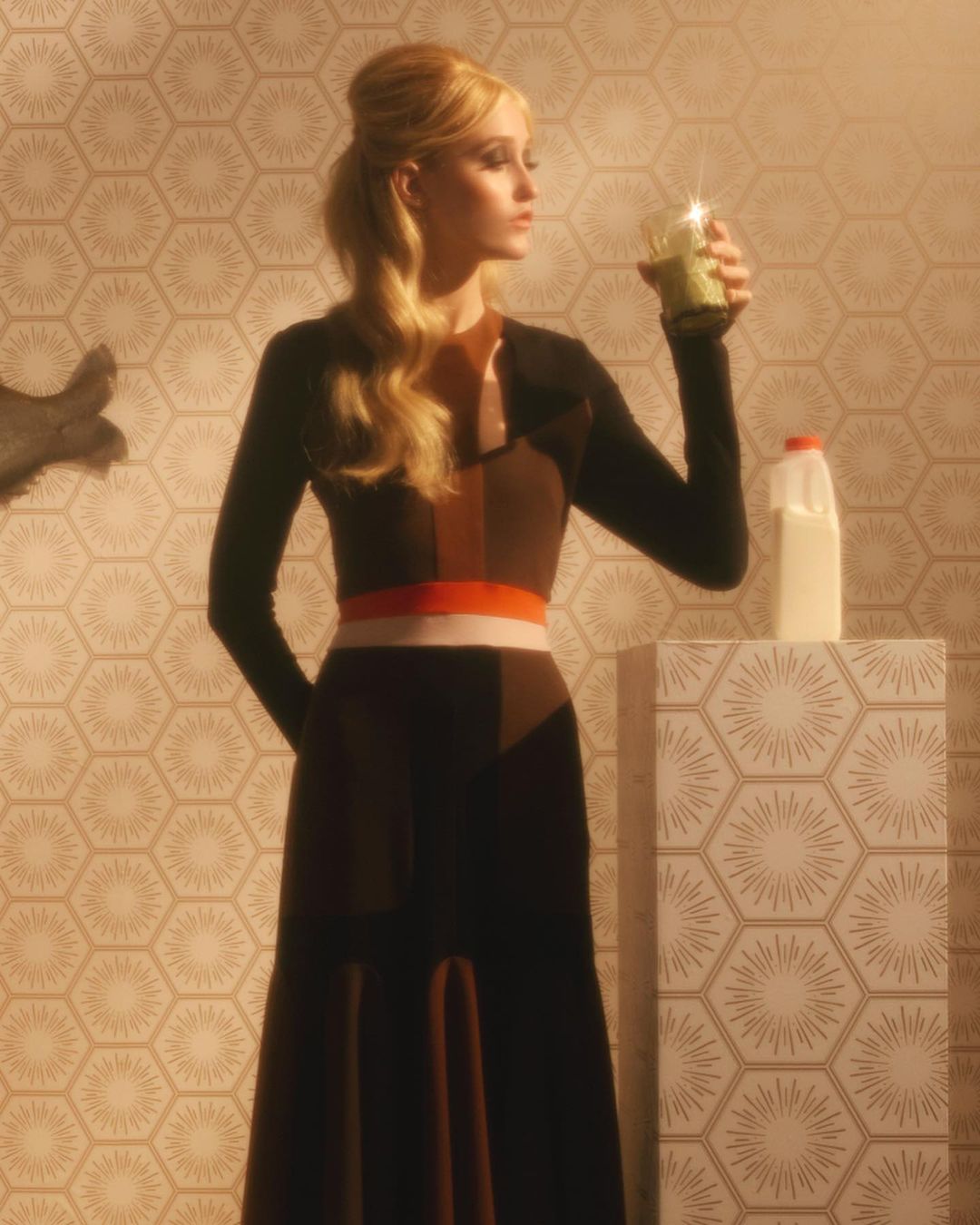 Woman poses indoors, holding a drink and wearing a long-sleeve midi dress, with a milk jug and patterned background.