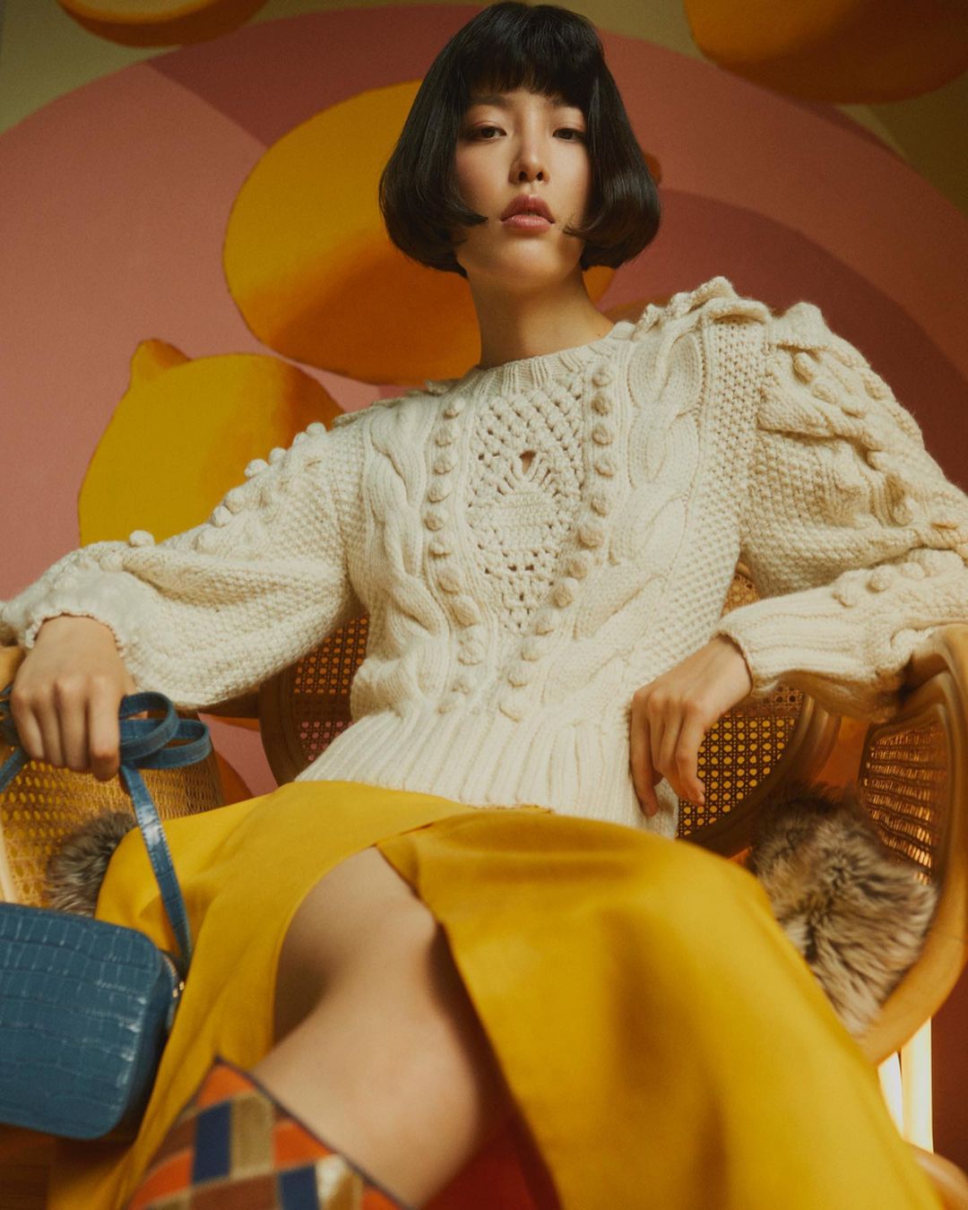 A fashionable Asian woman with a bob haircut sits in a wicker chair wearing a cream sweater, yellow skirt, boots, and blue handbag against lemon wallpaper.