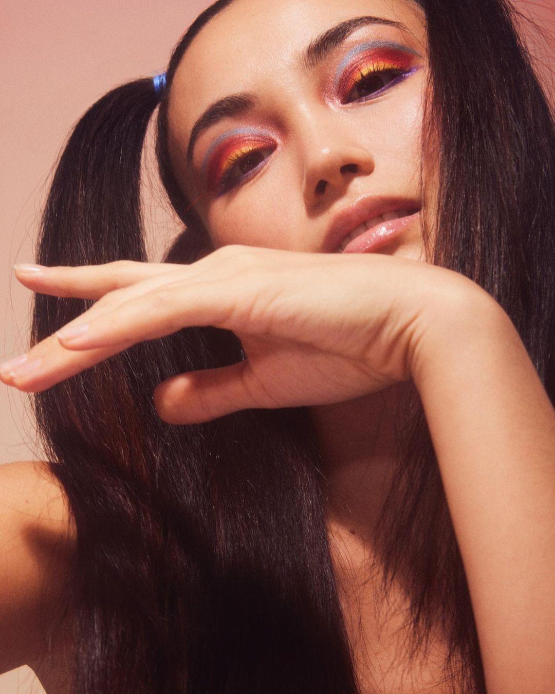 Close-up of an adult female with colorful eye makeup, styled hair, and fair skin, posing with a hand near her face against a pink background.