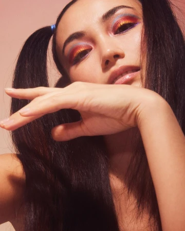 Close-up of an adult female with colorful eye makeup, styled hair, and fair skin, posing with a hand near her face against a pink background.