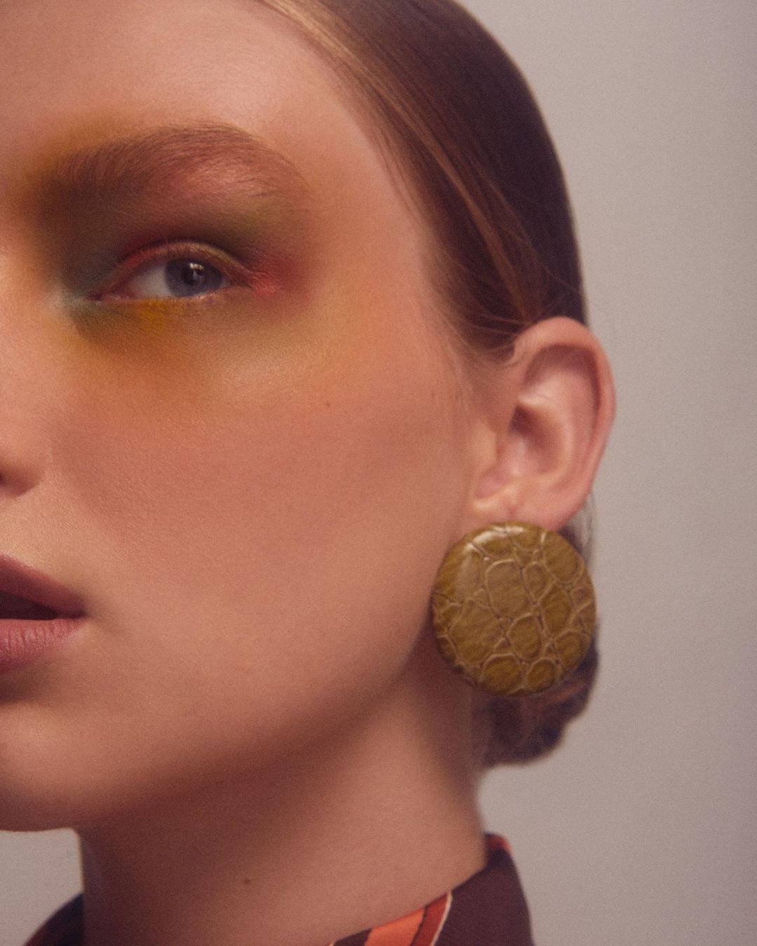 Close-up studio shot of a fair-skinned woman wearing colorful makeup and large disc earrings with a textured surface.