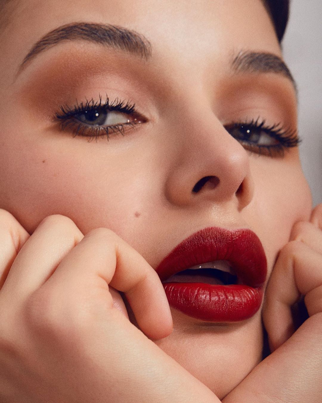 Close-up studio shot of a young adult woman with red lipstick, holding her hands near her face, soft lighting, beauty photography.