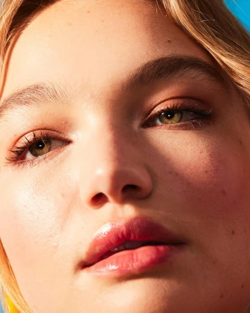 Close-up portrait of a young woman with blonde hair, green eyes, and glossy pink lips against a bright blue sky.