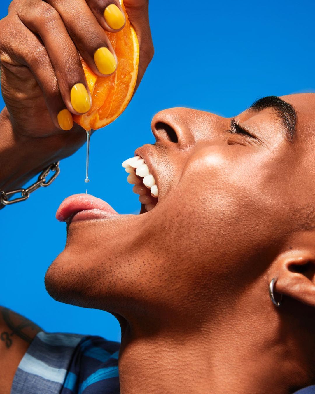 African-American man with open mouth catches dripping juice from an orange slice in a brightly lit studio shot against blue background.