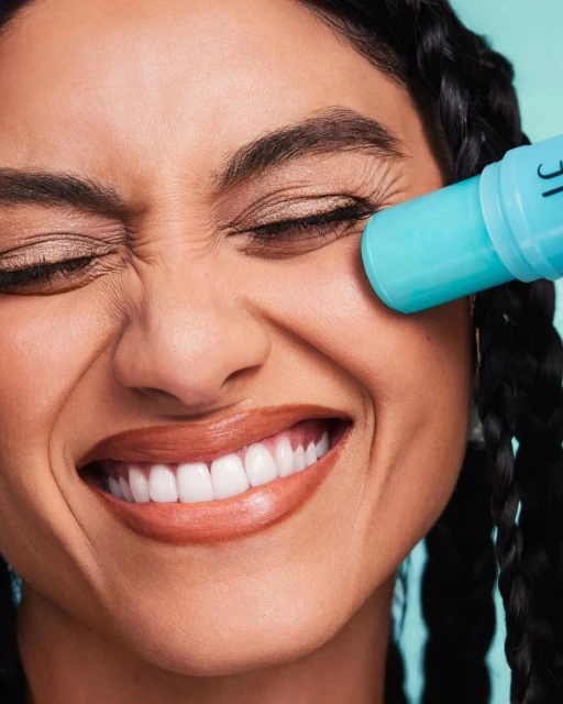 Close-up of a woman smiling with her eyes closed while applying a turquoise-colored beauty product to her face.